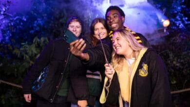 Four young adults dressed in Hogwarts-themed clothing smile and pose for a selfie in a magical, outdoor setting with atmospheric lighting and mist in the background.