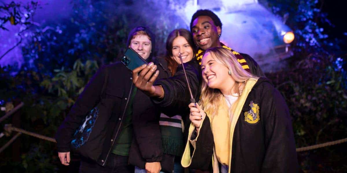 Four young adults dressed in Hogwarts-themed clothing smile and pose for a selfie in a magical, outdoor setting with atmospheric lighting and mist in the background.