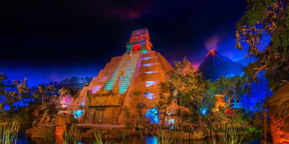 A brightly lit replica of a Mayan pyramid stands surrounded by lush greenery, with a glowing volcano erupting in the background under a dramatic, colorful night sky.
