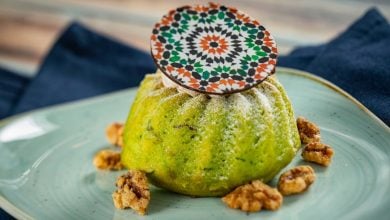 A vibrant green dessert dusted with powdered sugar, topped with a round patterned chocolate piece, is served on a light blue plate with walnuts scattered around. A dark blue cloth is in the background.