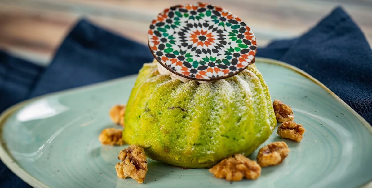 A vibrant green dessert dusted with powdered sugar, topped with a round patterned chocolate piece, is served on a light blue plate with walnuts scattered around. A dark blue cloth is in the background.