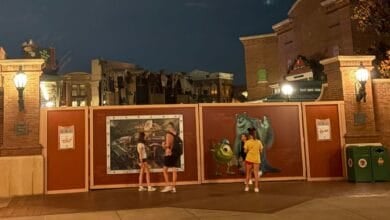 Three people stand in front of construction walls decorated with Monsters, Inc. characters Mike and Sulley at night in a theme park setting, with street lamps and brick buildings in the background.