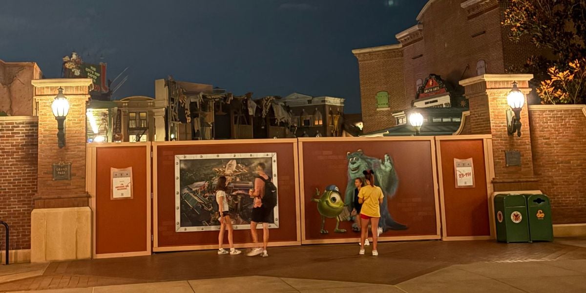 Three people stand in front of construction walls decorated with Monsters, Inc. characters Mike and Sulley at night in a theme park setting, with street lamps and brick buildings in the background.