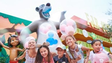 Five smiling children wearing Mickey and Minnie Mouse ears pose in front of large statues of Baloo and Mowgli from The Jungle Book at a colorful theme park, holding Mickey-shaped balloons.