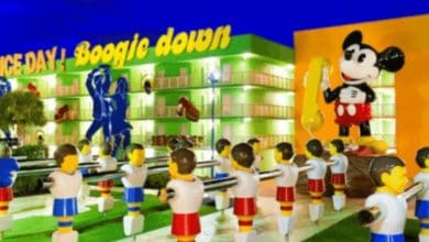 Colorful outdoor scene at Disney's Pop Century hotel with large foosball player statues, a giant cartoon mouse figure, and a bright green and yellow "GAME DAY! Boogie down" sign on a building in the background.