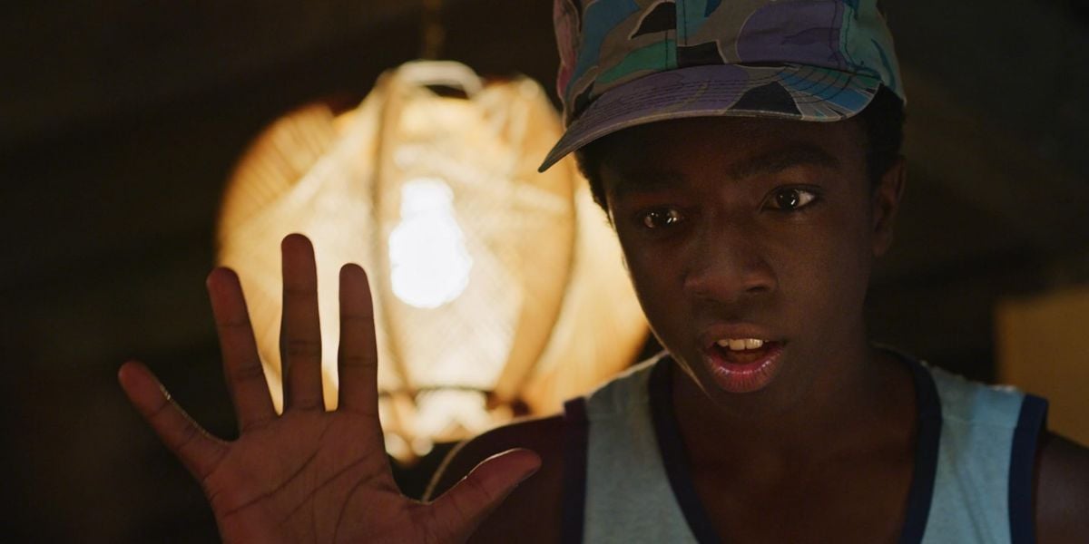 A boy wearing a colorful cap and tank top holds up his hand with fingers spread, looking surprised—like he’s just seen Disney magic. A bright, round light fixture glows in the background.