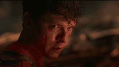 A young man in a red superhero suit, with cuts and blood on his face, looks intensely over his shoulder in a dark, smoky setting.