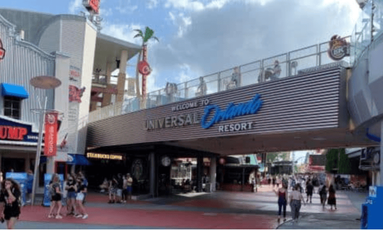 People walk beneath a large sign reading "Welcome to Universal Orlando Resort" at the entrance to Universal, with restaurants like Bubba Gump Shrimp Co. visible on either side.