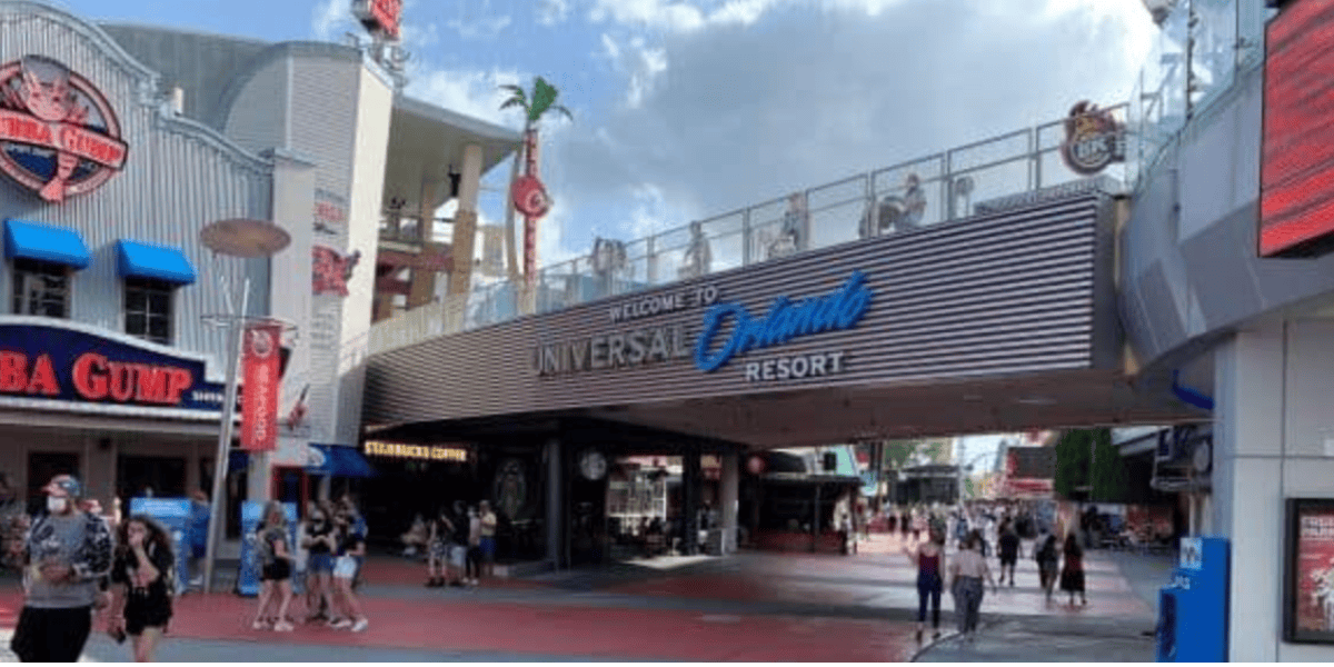 People walk beneath a large sign reading "Welcome to Universal Orlando Resort" at the entrance to Universal, with restaurants like Bubba Gump Shrimp Co. visible on either side.
