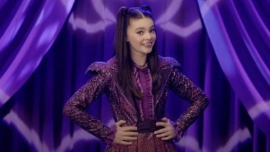 A young woman with long brown hair in pigtails, wearing a sparkly purple outfit, stands smiling with her hands on her hips in front of purple curtains with zigzag patterns.