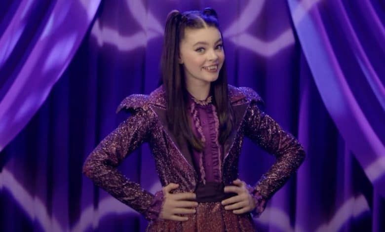 A young woman with long brown hair in pigtails, wearing a sparkly purple outfit, stands smiling with her hands on her hips in front of purple curtains with zigzag patterns.