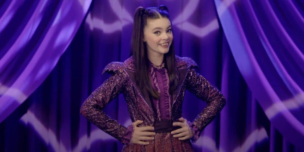 A young woman with long brown hair in pigtails, wearing a sparkly purple outfit, stands smiling with her hands on her hips in front of purple curtains with zigzag patterns.