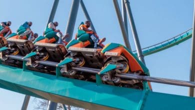 People riding a colorful roller coaster with orange and green cars, ascending a steep track with visible support beams, under a clear blue sky.