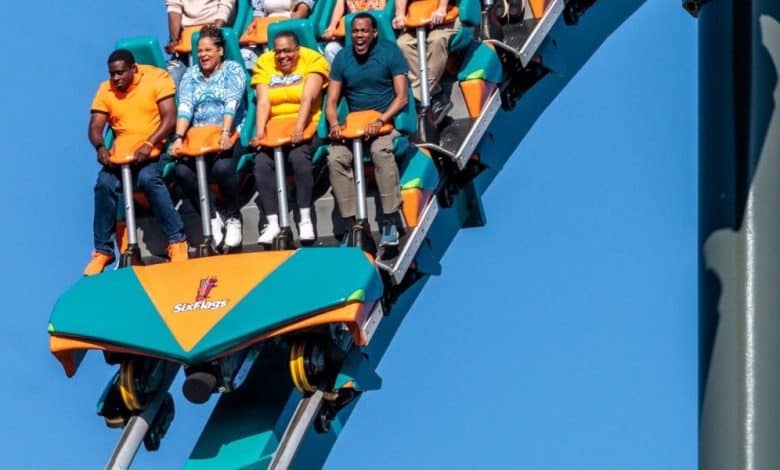 A group of people are riding a roller coaster at Six Flags, gripping the safety bars and expressing excitement and fear as the ride descends steeply under a clear blue sky.