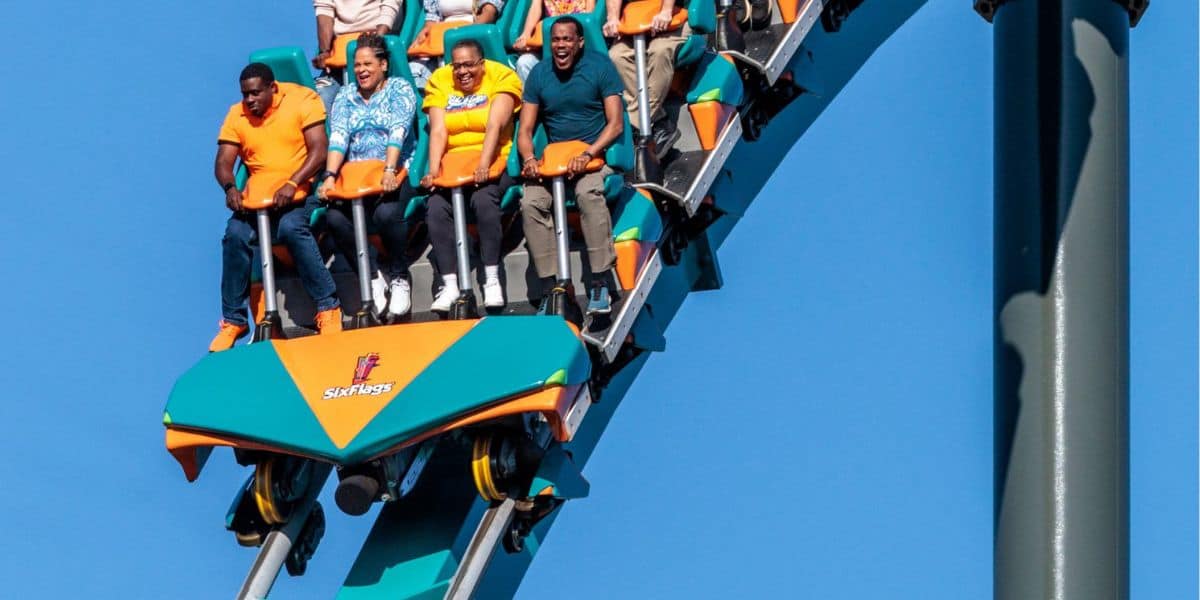 A group of people are riding a roller coaster at Six Flags, gripping the safety bars and expressing excitement and fear as the ride descends steeply under a clear blue sky.