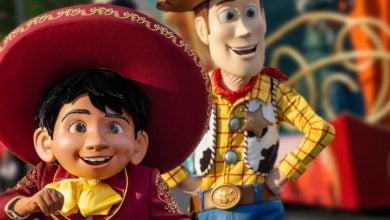 A costumed performer dressed as Miguel from "Coco" and another as Woody from "Toy Story" stand outdoors during a lively parade. Both characters are smiling and surrounded by festive decorations.