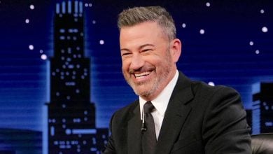 A man in a black suit and tie smiles while sitting at a desk on a talk show set, reminiscent of Jimmy Kimmel, with a city skyline and night sky backdrop behind him.