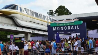 A large crowd waits beneath the Magic Kingdom Monorail sign as a monorail train passes overhead at a station, with trees and a partly cloudy sky in the background.