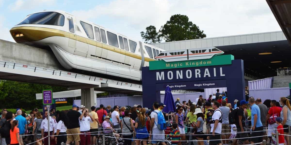 A large crowd waits beneath the Magic Kingdom Monorail sign as a monorail train passes overhead at a station, with trees and a partly cloudy sky in the background.
