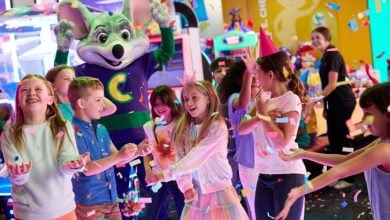 A group of kids celebrate at an arcade, smiling and throwing confetti. A person in a mouse mascot costume stands behind them with arms raised. Colorful lights and games fill the background.