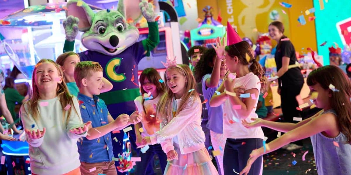 A group of kids celebrate at an arcade, smiling and throwing confetti. A person in a mouse mascot costume stands behind them with arms raised. Colorful lights and games fill the background.