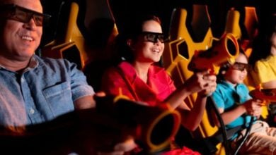 Four people wearing 3D glasses sit in yellow seats, smiling and holding toy blasters, appearing to enjoy an interactive ride or 3D attraction in a dark, amusement park-like setting.