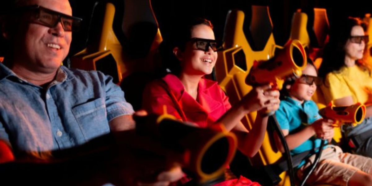 Four people wearing 3D glasses sit in yellow seats, smiling and holding toy blasters, appearing to enjoy an interactive ride or 3D attraction in a dark, amusement park-like setting.