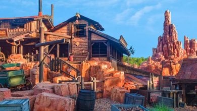 A rustic, Old West-themed roller coaster station with wooden buildings, mining props, and red rocky terrain, set against a blue sky and a jagged mountain peak in the background.