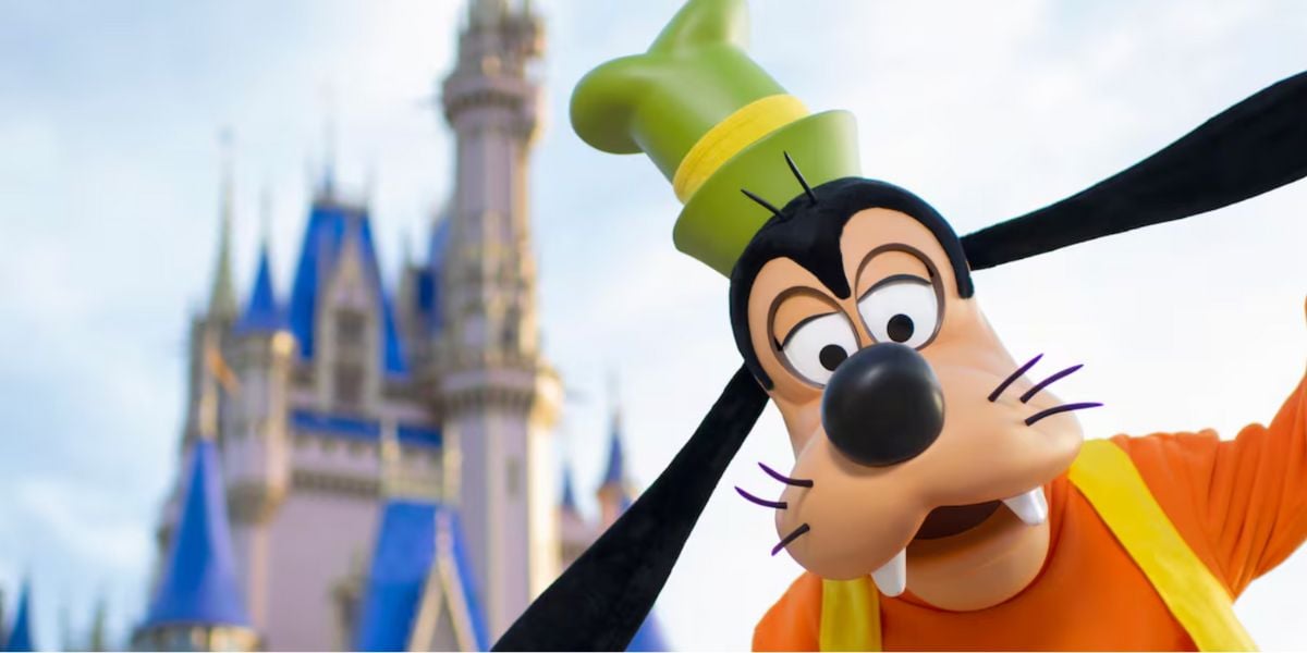 Goofy, a Disney character in an orange shirt and green hat, leans in playfully toward the camera with a blurred Cinderella Castle in the background at a Disney theme park.