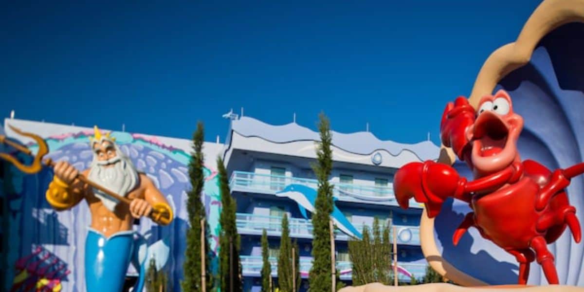 Large statues of King Triton and Sebastian the crab from "The Little Mermaid" stand outside a colorful, ocean-themed building under a clear blue sky.