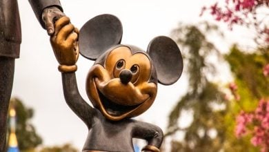 A bronze statue of Mickey Mouse smiling and holding hands with a larger figure, with trees and pink flowers in the blurred background.