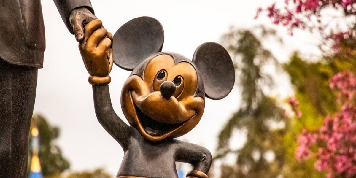 A bronze statue of Mickey Mouse smiling and holding hands with a larger figure, with trees and pink flowers in the blurred background.