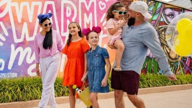 A Disney-loving family of five, including three children wearing mouse ears, walks happily in front of a colorful mural that reads "WARM." The man holds a Mickey balloon, and everyone is smiling in bright, casual clothing.