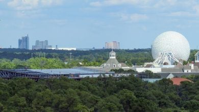 A distant view of Epcot’s Spaceship Earth, surrounded by trees and buildings, with a city skyline and a yellow arrow pointing to a tall building in the far background under a partly cloudy sky.