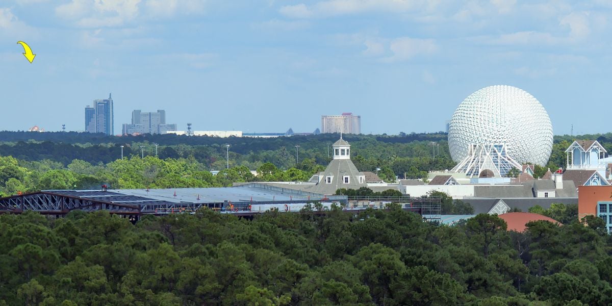 A distant view of Epcot’s Spaceship Earth, surrounded by trees and buildings, with a city skyline and a yellow arrow pointing to a tall building in the far background under a partly cloudy sky.