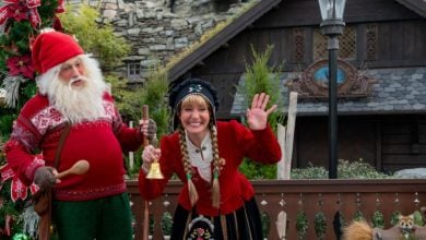 A man dressed as a traditional Santa and a woman in festive Scandinavian attire stand smiling and waving by a Christmas tree in front of a rustic wooden building, creating a classic Christmas scene at EPCOT.