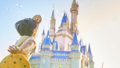A young child wearing a yellow dress and Mickey Mouse ears reaches out joyfully toward a fairytale castle with blue rooftops under a bright, sunny sky.