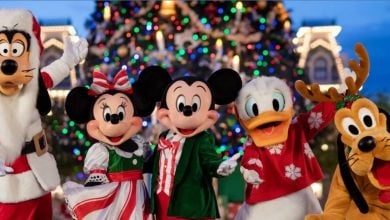 Goofy, Minnie Mouse, Mickey Mouse, Donald Duck, and Pluto dressed in festive holiday outfits stand in front of a brightly lit Christmas tree, smiling and posing for a cheerful holiday photo.
