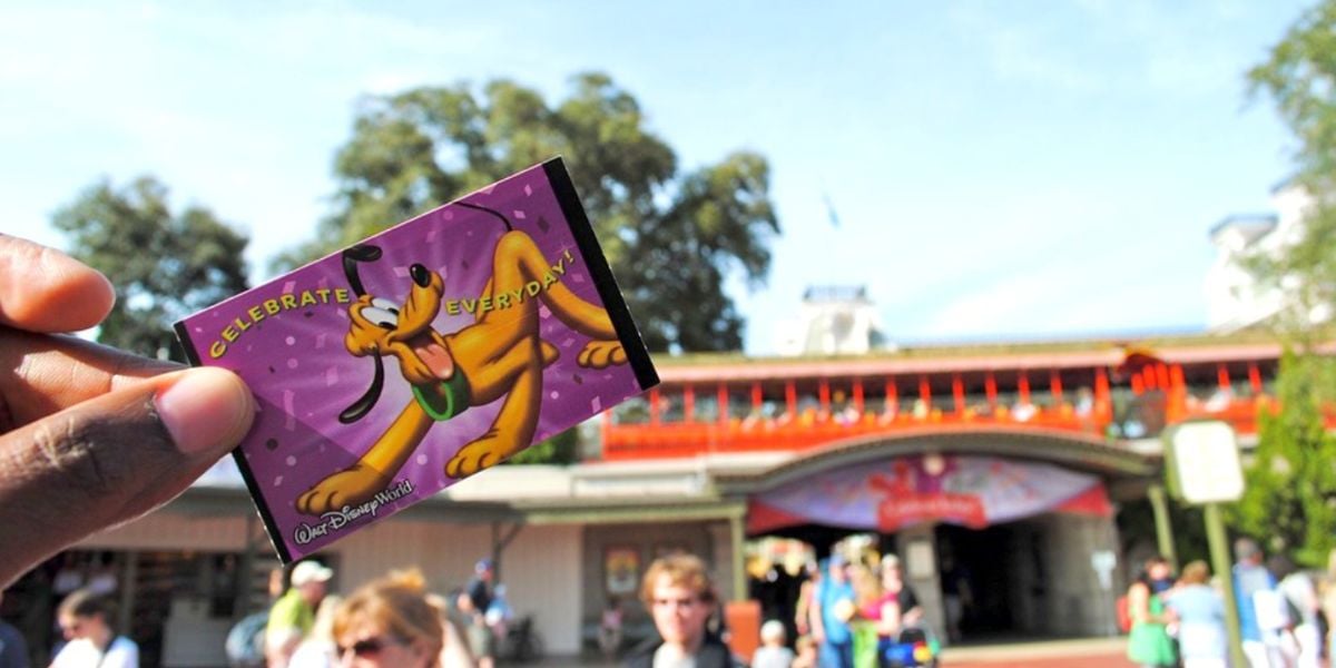 A hand holds a Disney World ticket featuring Pluto in front of the Magic Kingdom Park entrance. This is an old style of paper Disney World tickets.