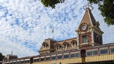 A brick building with a clock tower is decorated with festive garlands, set against a blue sky with scattered clouds. The roof has ornate detailing, and part of a train car is visible below the clock. Tree branches frame the top corners.