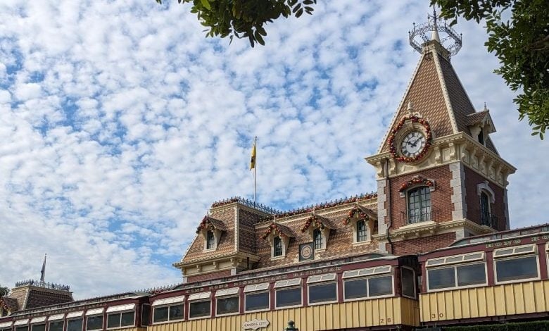 A brick building with a clock tower is decorated with festive garlands, set against a blue sky with scattered clouds. The roof has ornate detailing, and part of a train car is visible below the clock. Tree branches frame the top corners.