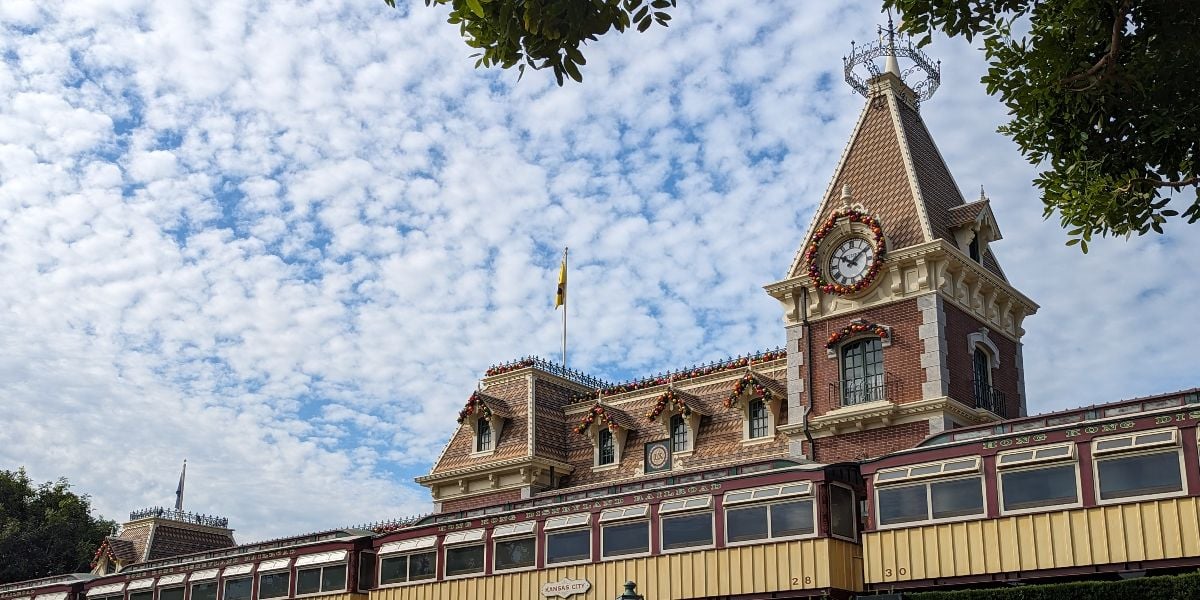 A brick building with a clock tower is decorated with festive garlands, set against a blue sky with scattered clouds. The roof has ornate detailing, and part of a train car is visible below the clock. Tree branches frame the top corners.
