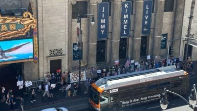 A crowd of protesters holding signs stands outside the "Jimmy Kimmel Live" studio in Hollywood, near a Metro bus and the El Capitan Theatre, seen from above.