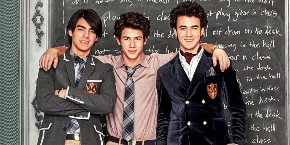 Three young men in school uniforms stand together in front of a chalkboard, smiling with their arms around each other. The chalkboard has handwritten text covering it.