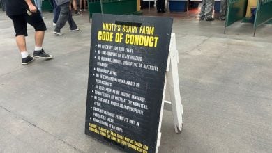 A black sandwich board sign displays the Knott’s Scary Farm Code of Conduct, listing rules for guest behavior. People are seen walking nearby on a concrete surface.