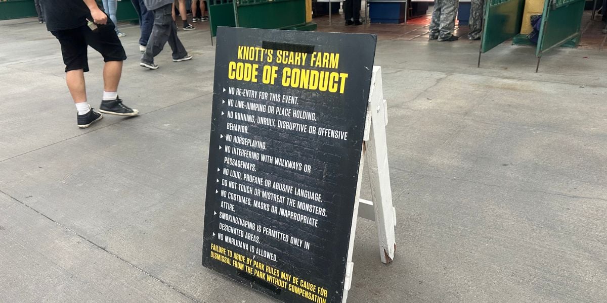 A black sandwich board sign displays the Knott’s Scary Farm Code of Conduct, listing rules for guest behavior. People are seen walking nearby on a concrete surface.