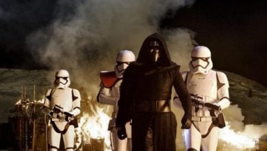 A hooded figure, Kylo Ren, in black robes stands in front of two armored soldiers in white, with a fiery explosion and smoke illuminating the snowy landscape at night.
