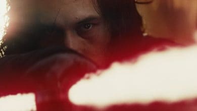 A close-up of a person with intense eyes and long hair holding a glowing red lightsaber, partially obscured by a blurred red light in the foreground.
