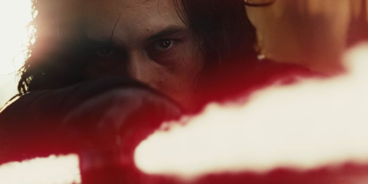 A close-up of a person with intense eyes and long hair holding a glowing red lightsaber, partially obscured by a blurred red light in the foreground.