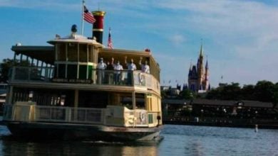 A ferryboat named "Admiral Joe Fowler" crosses a lake with passengers on board, American flags flying, and Cinderella Castle visible in the background under a blue sky.